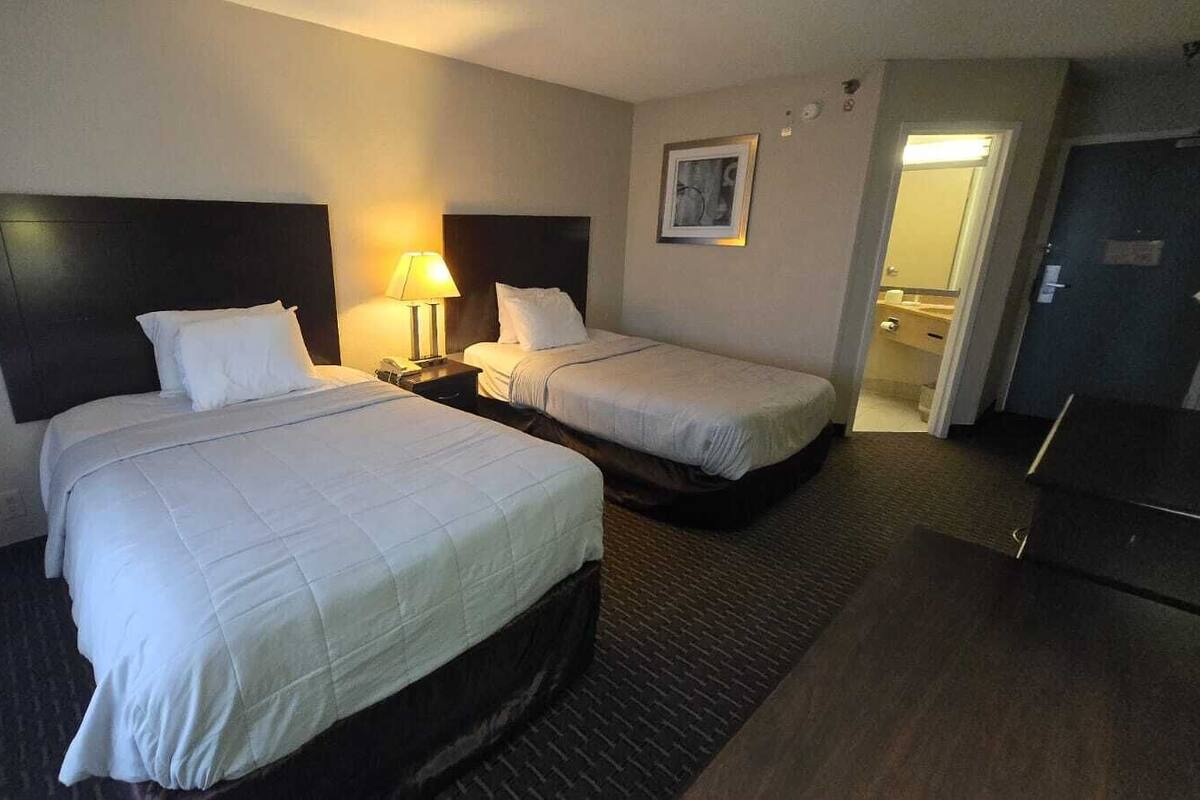 Une chambre d’hôtel avec deux lits, un bureau, une chaise, une lampe et une porte donnant sur une salle de bain.
