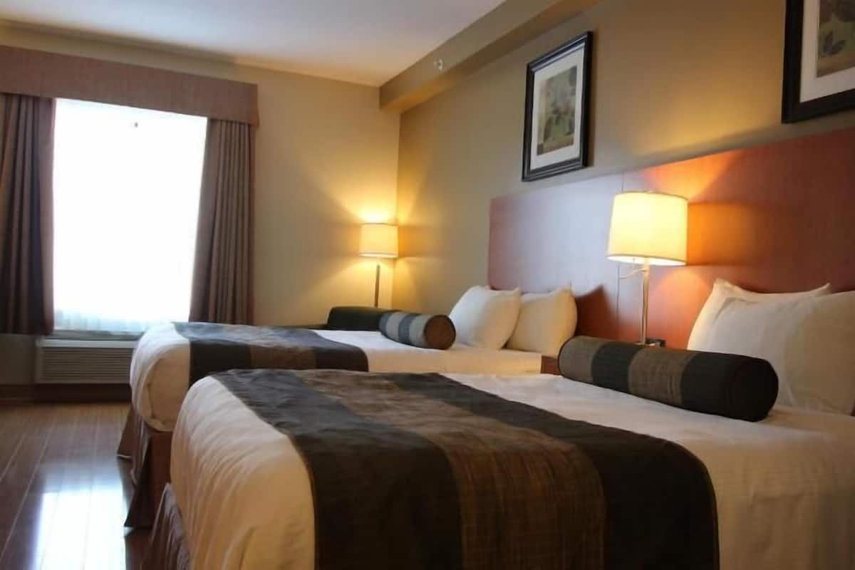 Une chambre d’hôtel avec deux lits, une grande fenêtre avec des rideaux, deux lampes fixées au mur et des tableaux encadrés accrochés au mur.