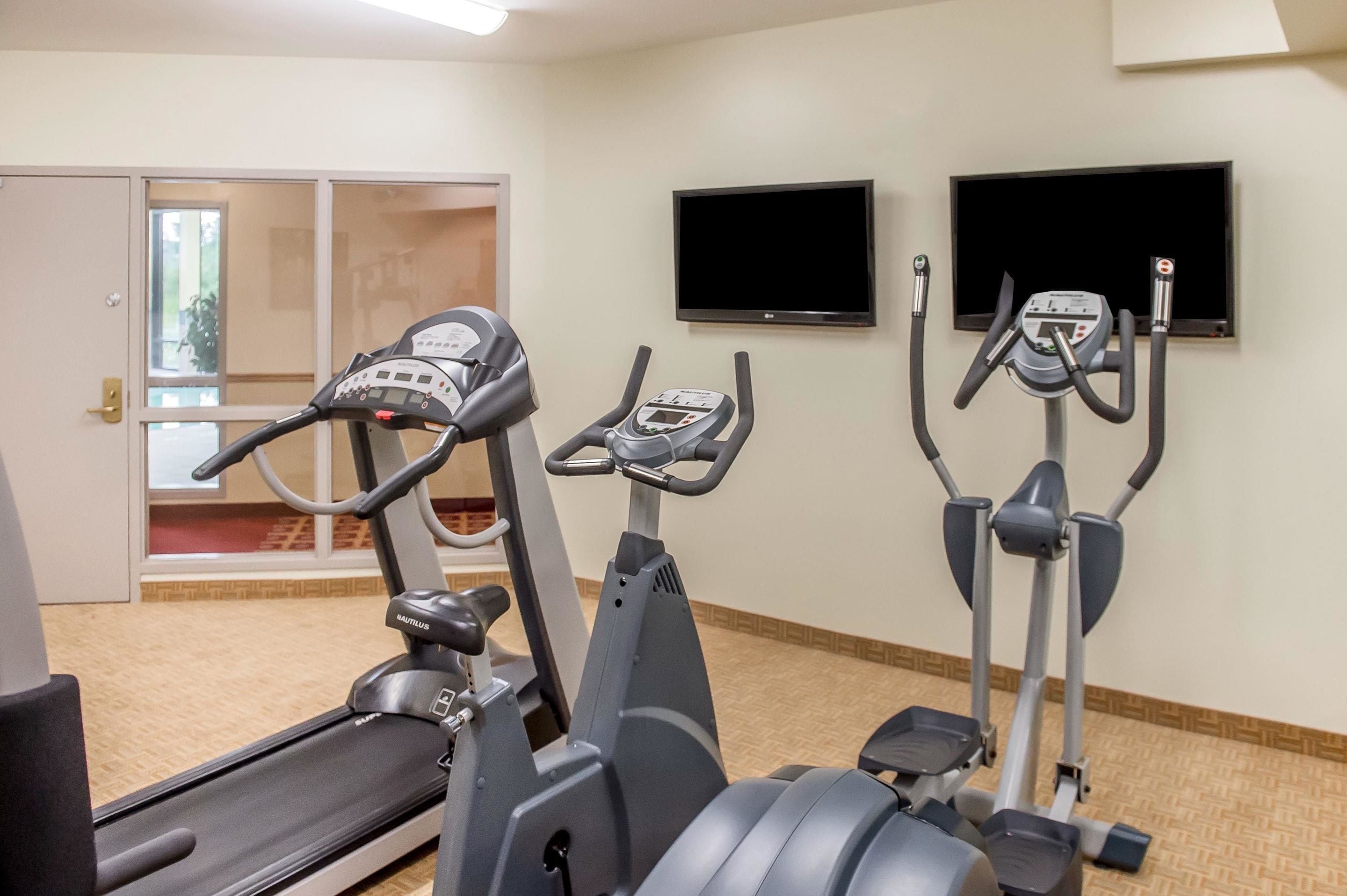 A gym with treadmills, an elliptical, and a stationary bike. There is a TV mounted on the wall and a door leading outside.