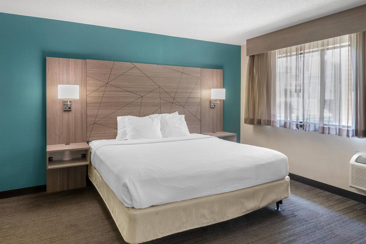 Quarto de hotel com uma cama grande, duas mesas de cabeceira, cabeceira com padrão geométrico e uma janela com cortinas translúcidas.