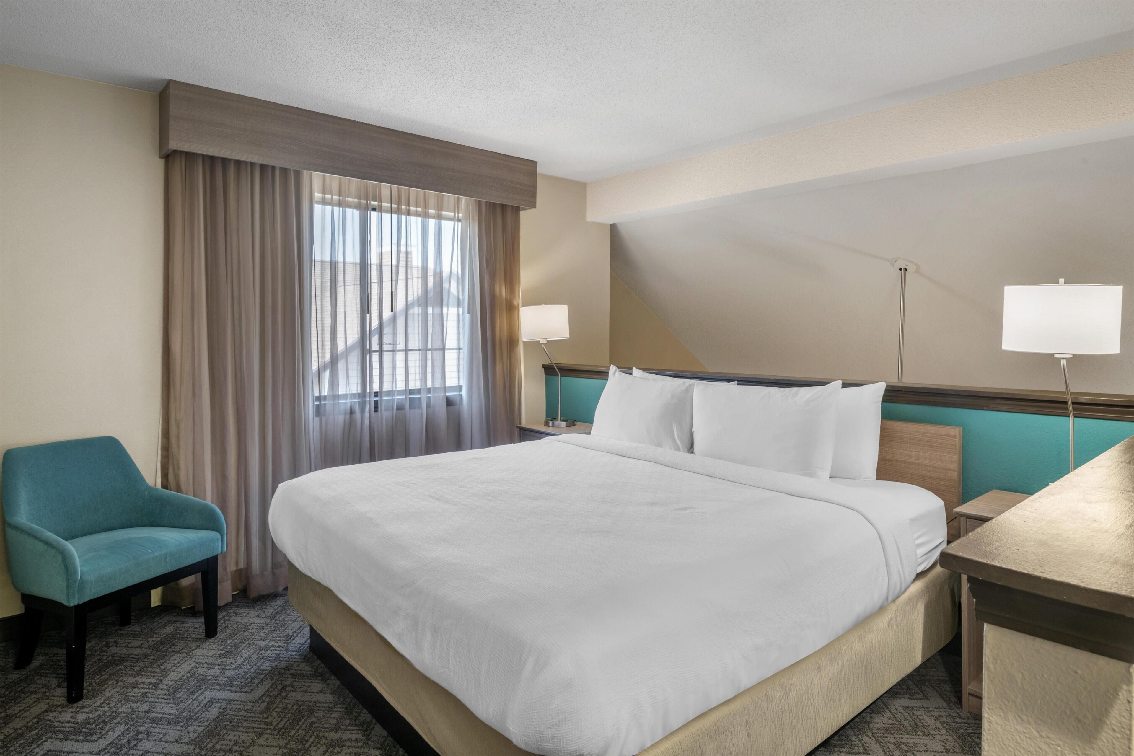 A hotel room with a bed, a chair, a desk, a lamp, and a window with curtains.
