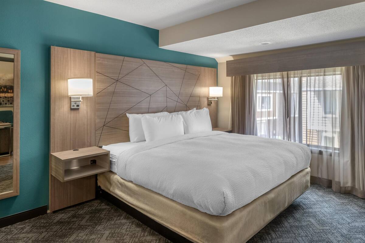 Quarto de hotel com cama grande, mesa de cabeceira, luminária na parede e janela com cortinas.