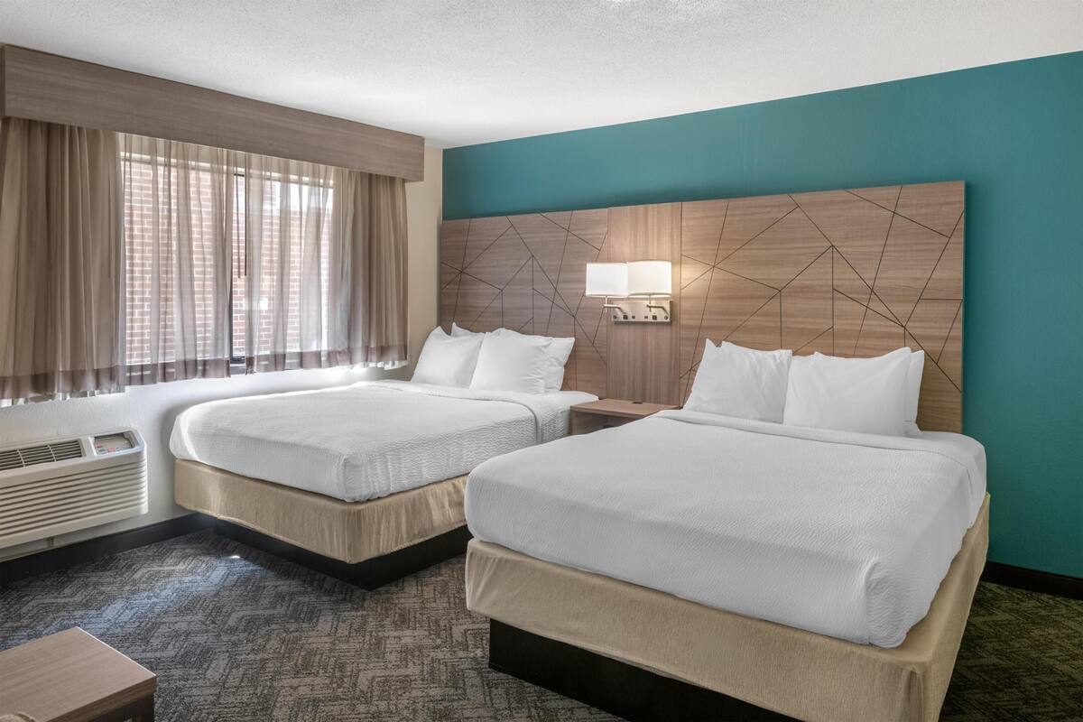 Quarto de hotel com duas camas, uma janela com cortinas, luminária na parede e um aquecedor.