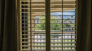 Una ventana con persianas horizontales, vista a un balcón con follaje y montañas al fondo.
