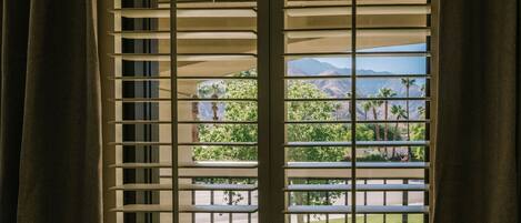 Una ventana con persianas horizontales, vista a un balcón con follaje y montañas al fondo.