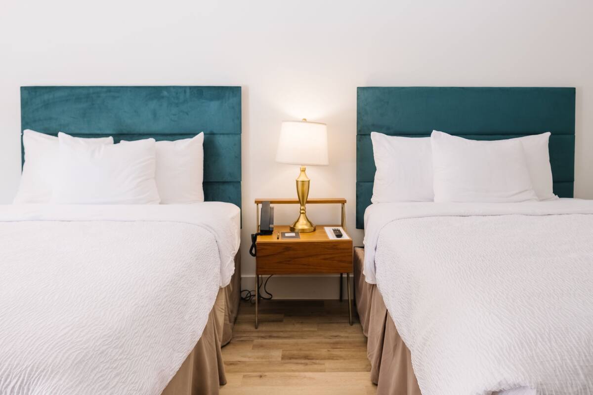 A hotel room with two beds, each with white bedding and teal headboards. There is a wooden nightstand between the beds with a gold lamp and a phone.