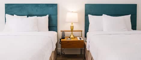 A hotel room with two beds, each with white bedding and teal headboards. There is a wooden nightstand between the beds with a gold lamp and a phone.