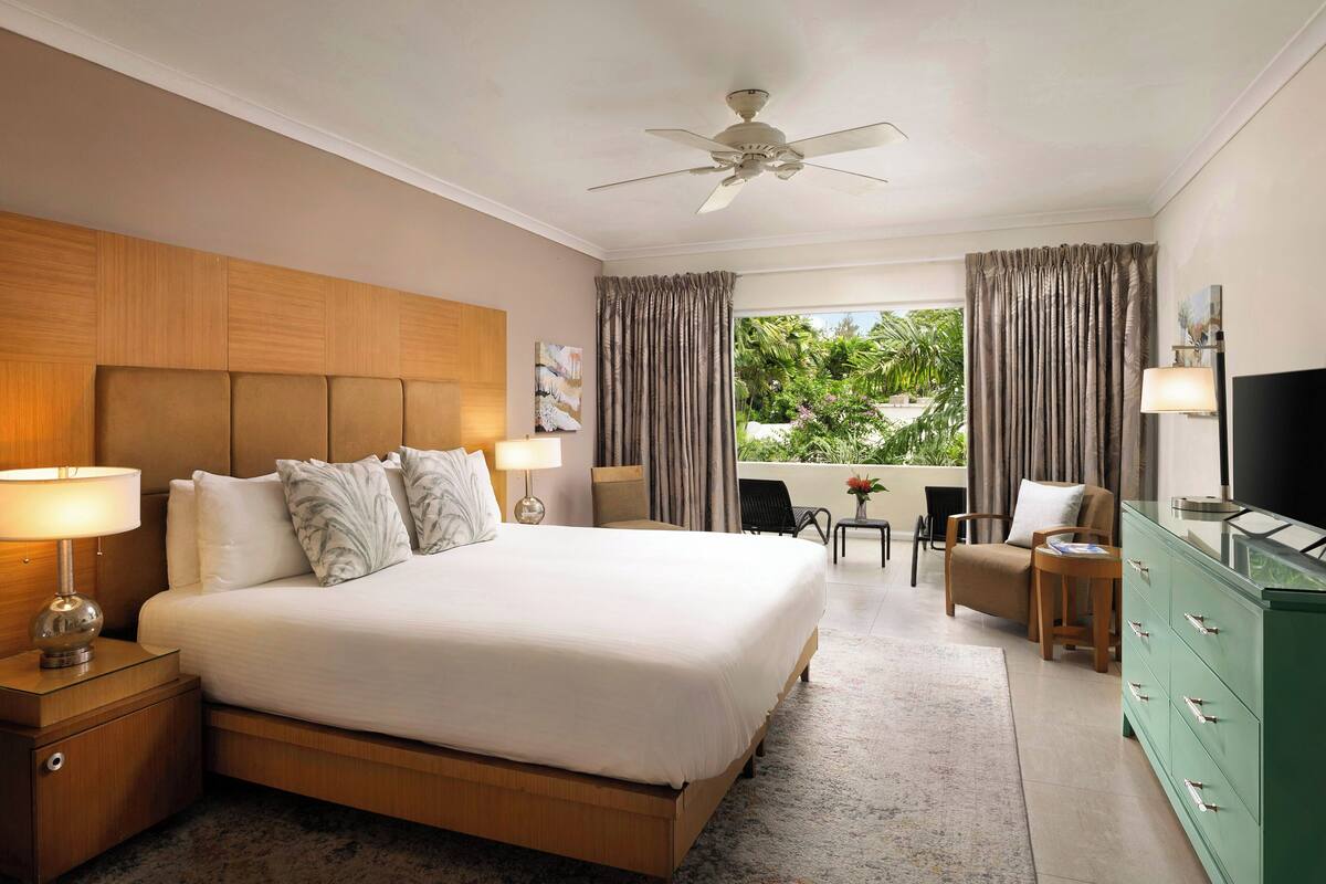 A hotel room with a large bed, a ceiling fan, a TV, a green dresser, and a balcony with curtains.