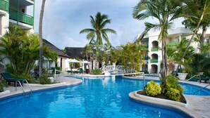 A swimming pool surrounded by palm trees and lounge chairs, with a building featuring balconies in the background. - Mango Bay All Inclusive (Holetown)