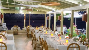 A covered outdoor dining area with tables set for a meal, chairs, and a view of the sea. - Mango Bay All Inclusive (Holetown)