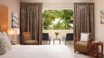 A hotel room with a large bed, two armchairs, a small table with a vase of flowers, and a view of a tropical landscape through sheer curtains.