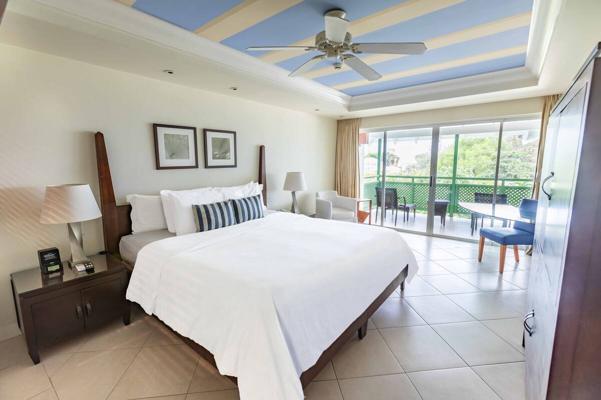 A hotel room with a large bed, a nightstand with a lamp, a ceiling fan, and a balcony with outdoor furniture.