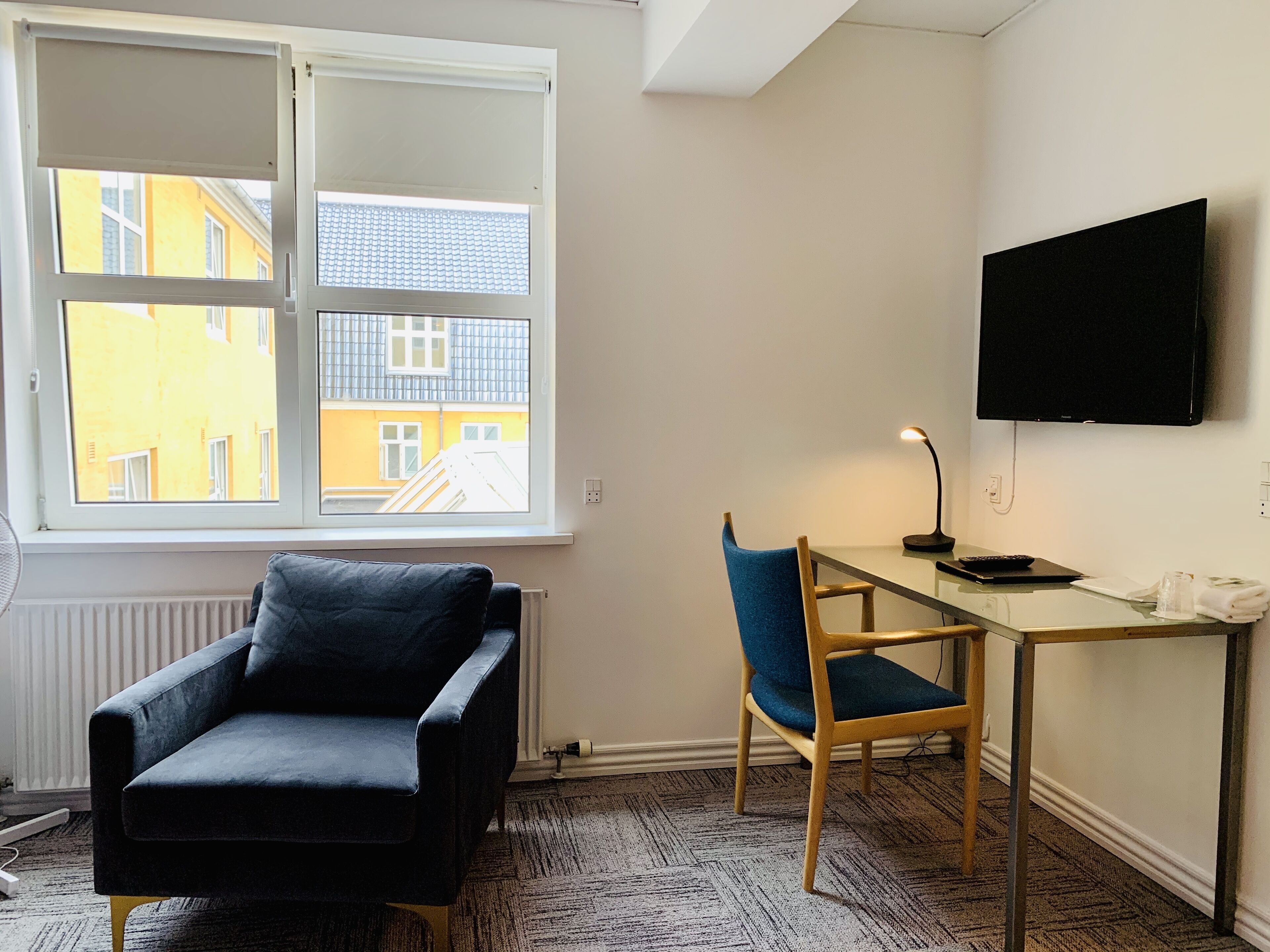 Una habitación con una silla negra, una silla de madera, un escritorio con una lámpara y un televisor en la pared.