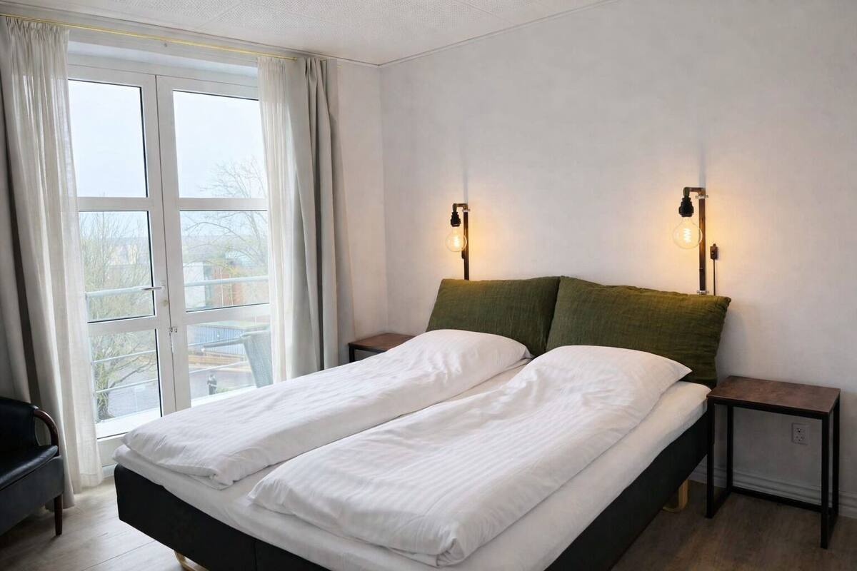 Een tweepersoonsbed met witte lakens, twee nachtkastjes en twee wandlampen.