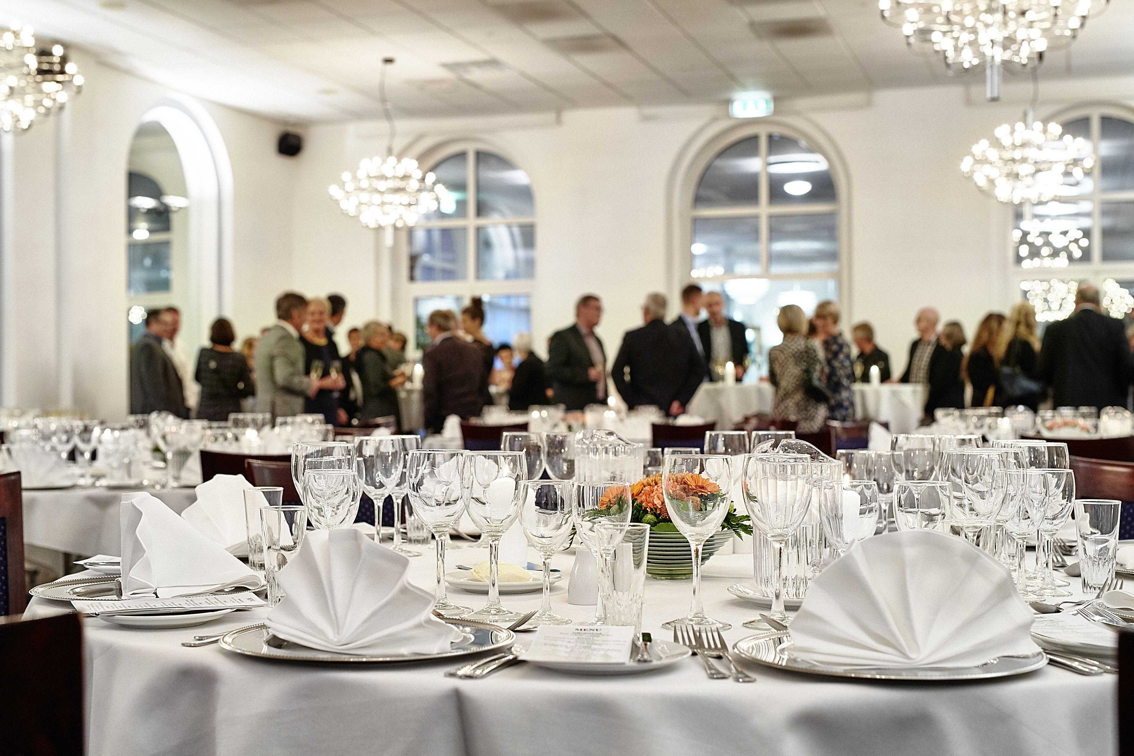 A formal dining room with tables set for a meal, large windows, and chandeliers.