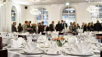 A formal dining room with tables set for a meal, large windows, and chandeliers.