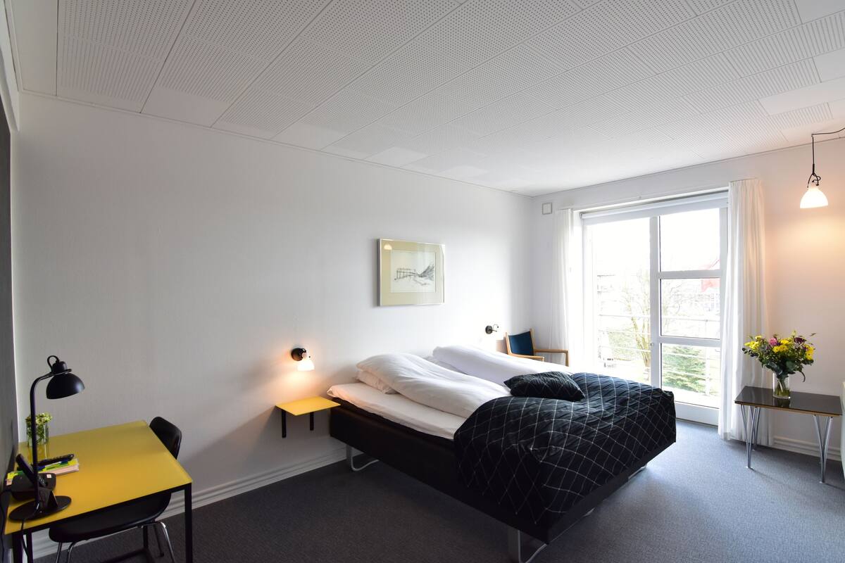 Een moderne hotelkamer met een bed, een bureau, een stoel, een klein tafeltje met een lamp en een raam met gordijnen.