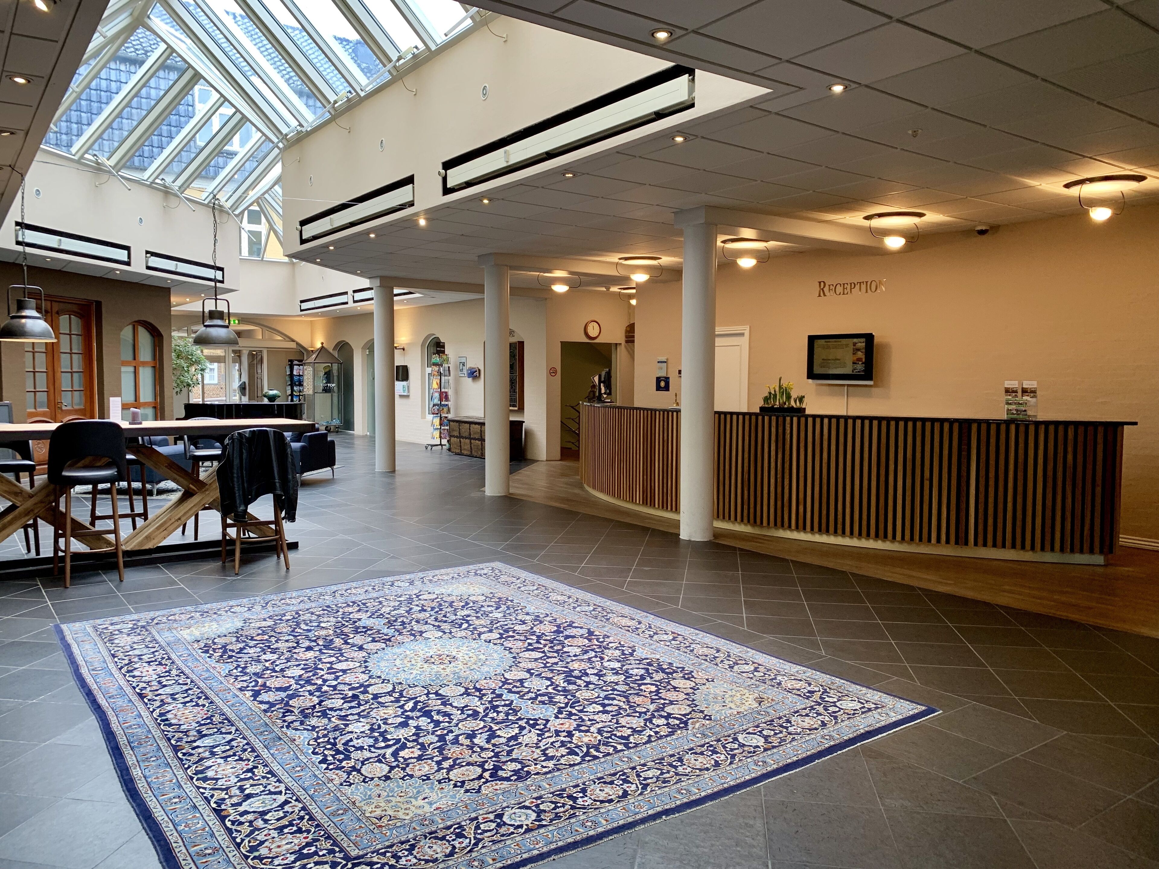 En rummelig lobby med en receptionsdisk, en stor gulvtæppe med et mønster, og et ovenlys.