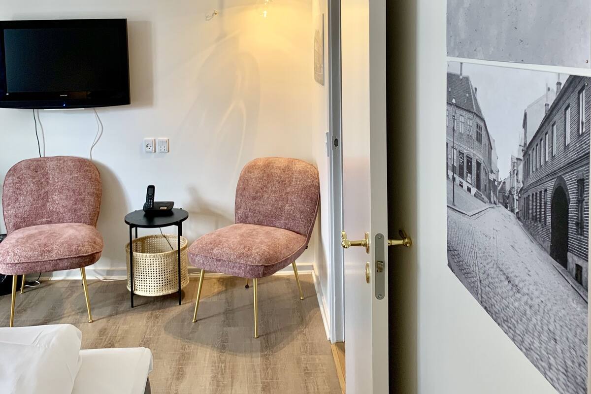 Een moderne hotelkamer met een flatscreen televisie, twee roze fauteuils, een klein bijzettafeltje met een mand, en een grote zwart-wit foto aan de muur.