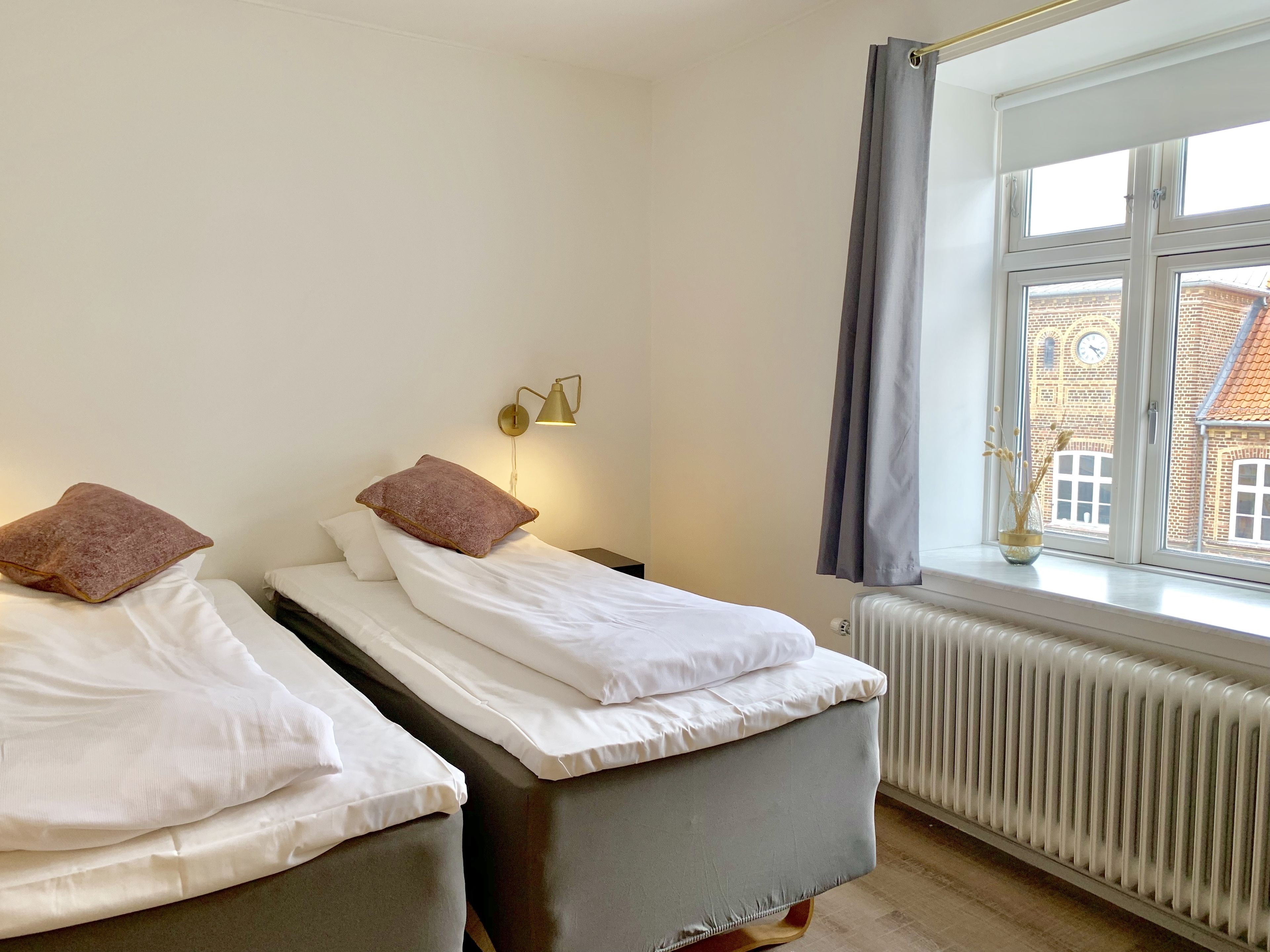 A room with two beds, a radiator, a window with curtains, and a lamp.