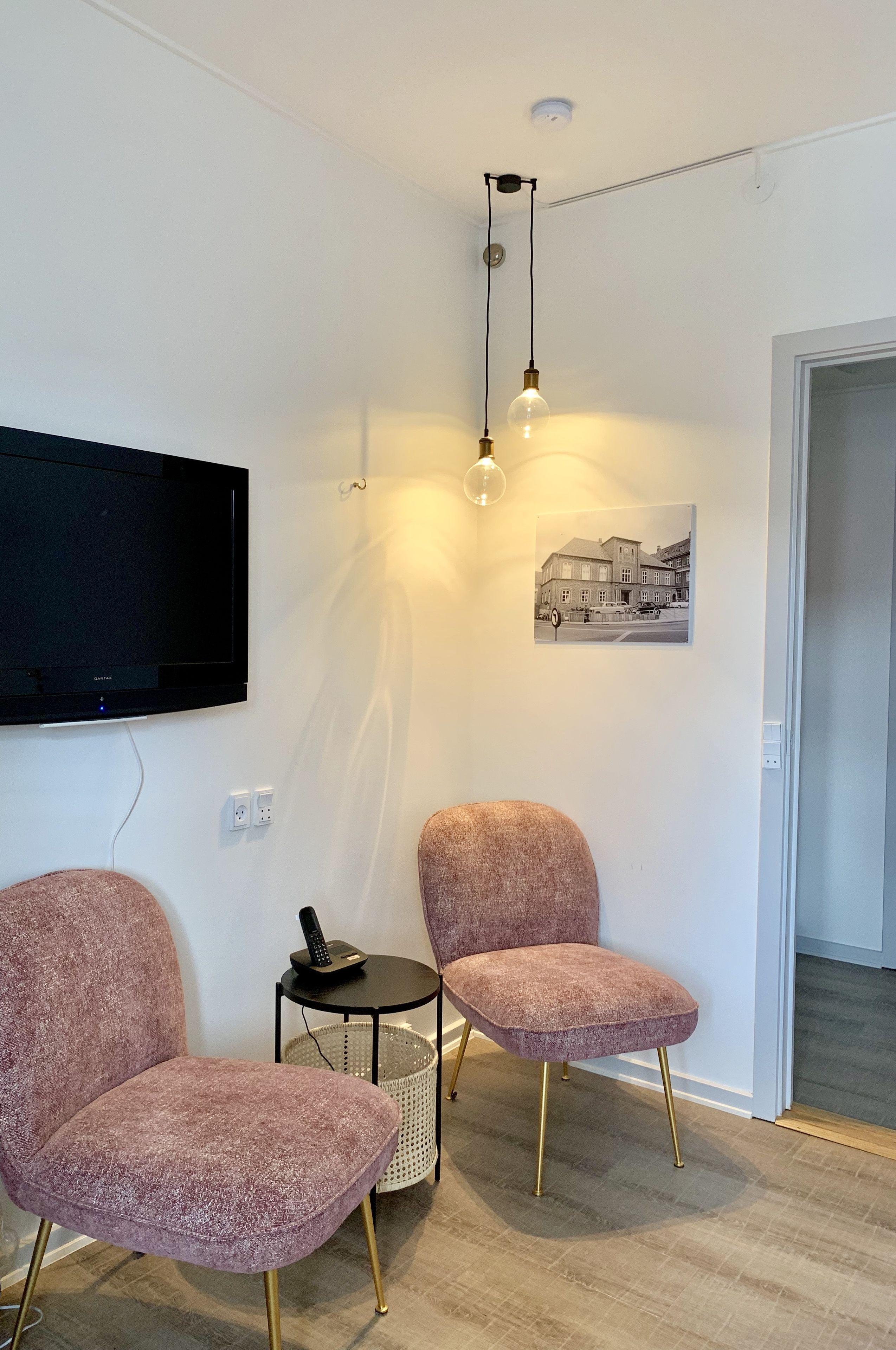 A modern room with a flat-screen TV, two pink chairs, a small round table with a basket, and a hanging light fixture.