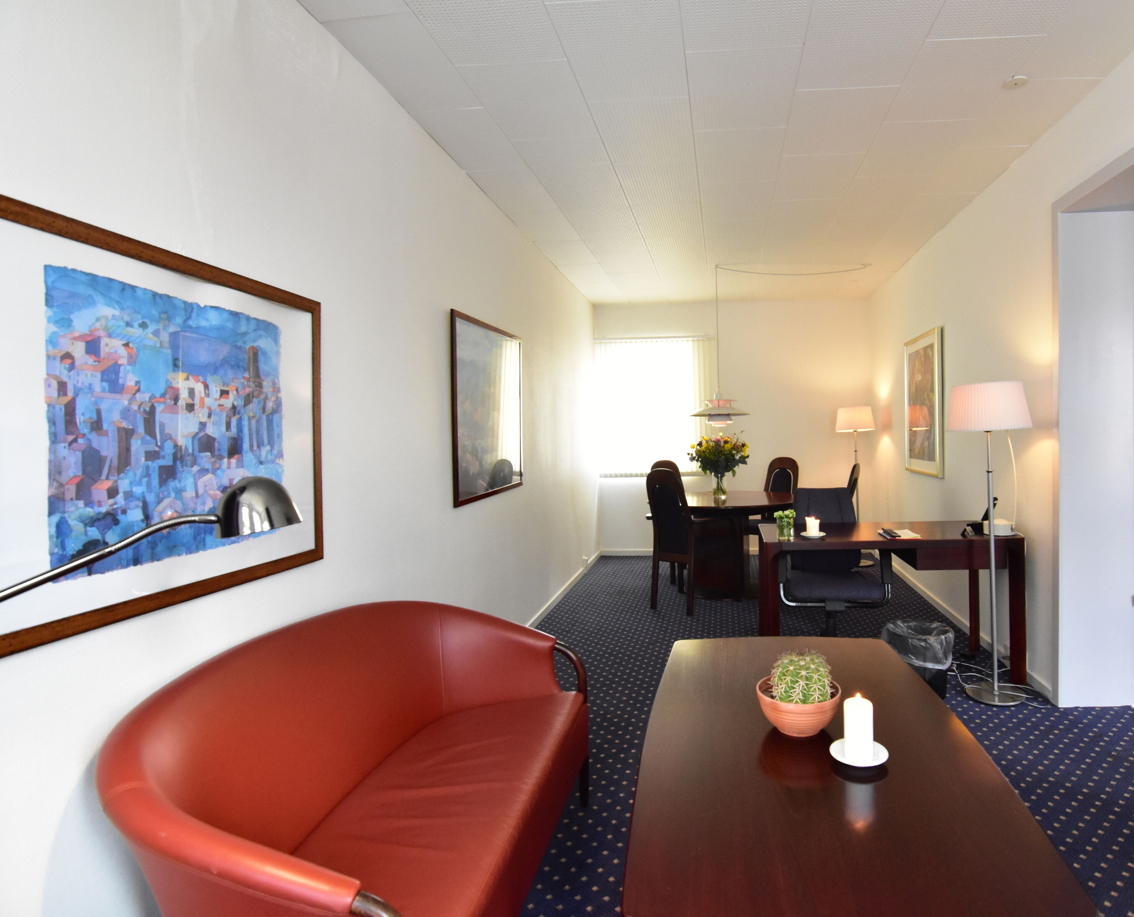 Habitación de hotel con sofá de cuero rojo, una mesa de comedor con sillas, un pequeño cactus en la mesa y un cuadro en la pared.