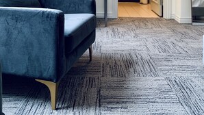Een moderne hotelkamer met een grote flatscreen televisie aan de muur, een comfortabele donkerblauwe bank, een klein bijzettafeltje met een lamp, en uitzicht op de badkamer door een geopende deur.