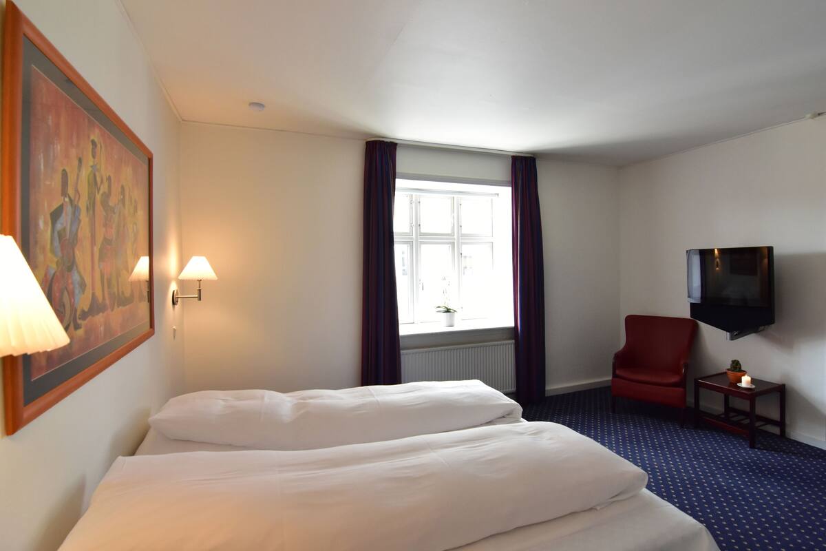 Een hotelkamer met twee bedden, een rode fauteuil, een klein tafeltje met een plant, een schilderij aan de muur, en een aan de muur bevestigde televisie.