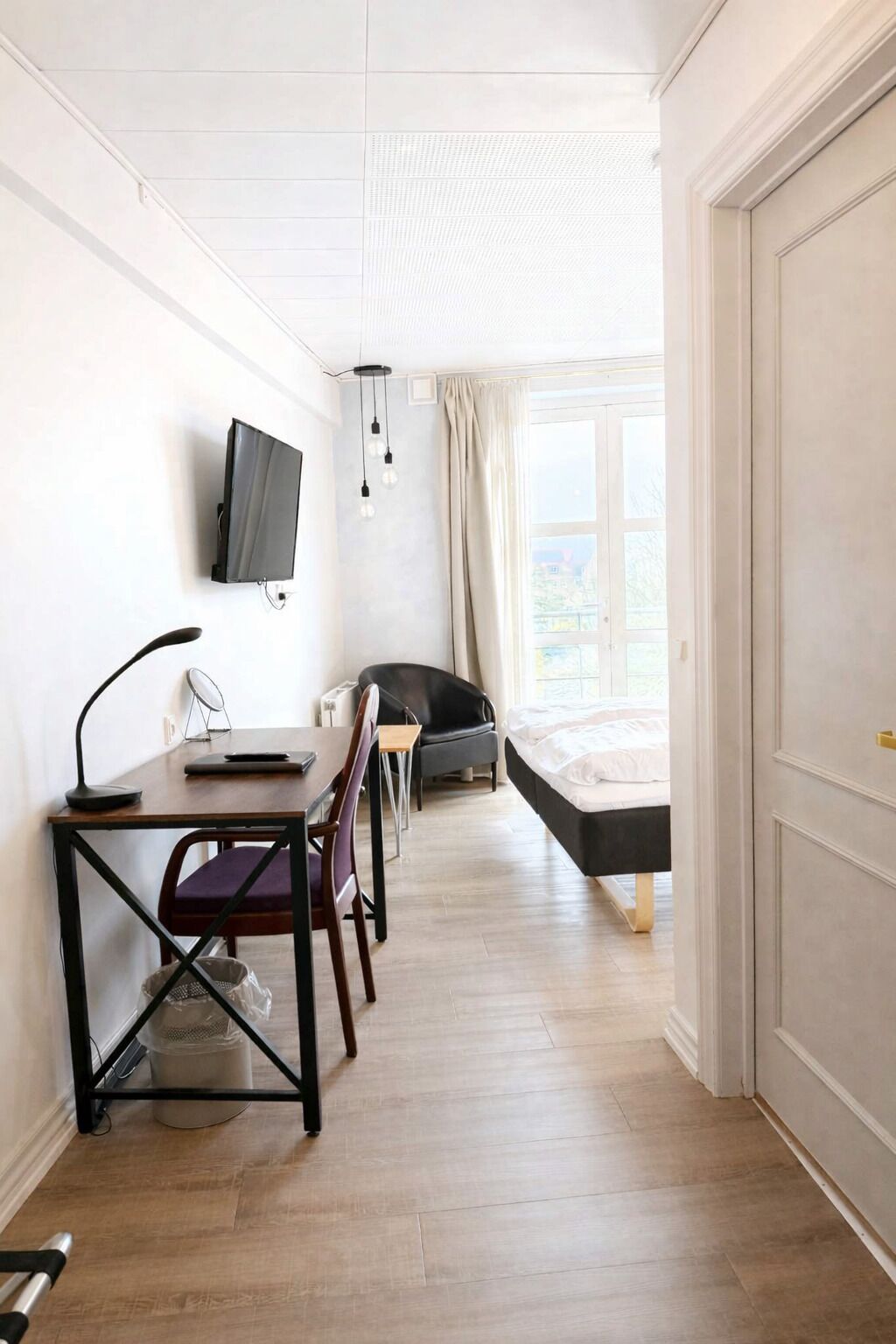 Una habitación con una cama, un escritorio con una silla, un televisor montado en la pared y una ventana con cortinas.