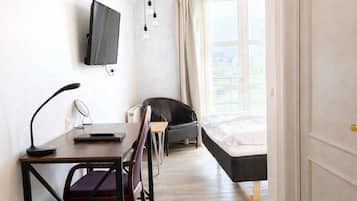 Una habitación con una cama, un escritorio con una silla, un televisor montado en la pared y una ventana con cortinas.