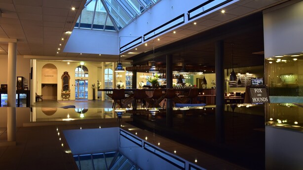 Een ruime atrium met glazen dak, een bar en een zithoek met tafels en stoelen.