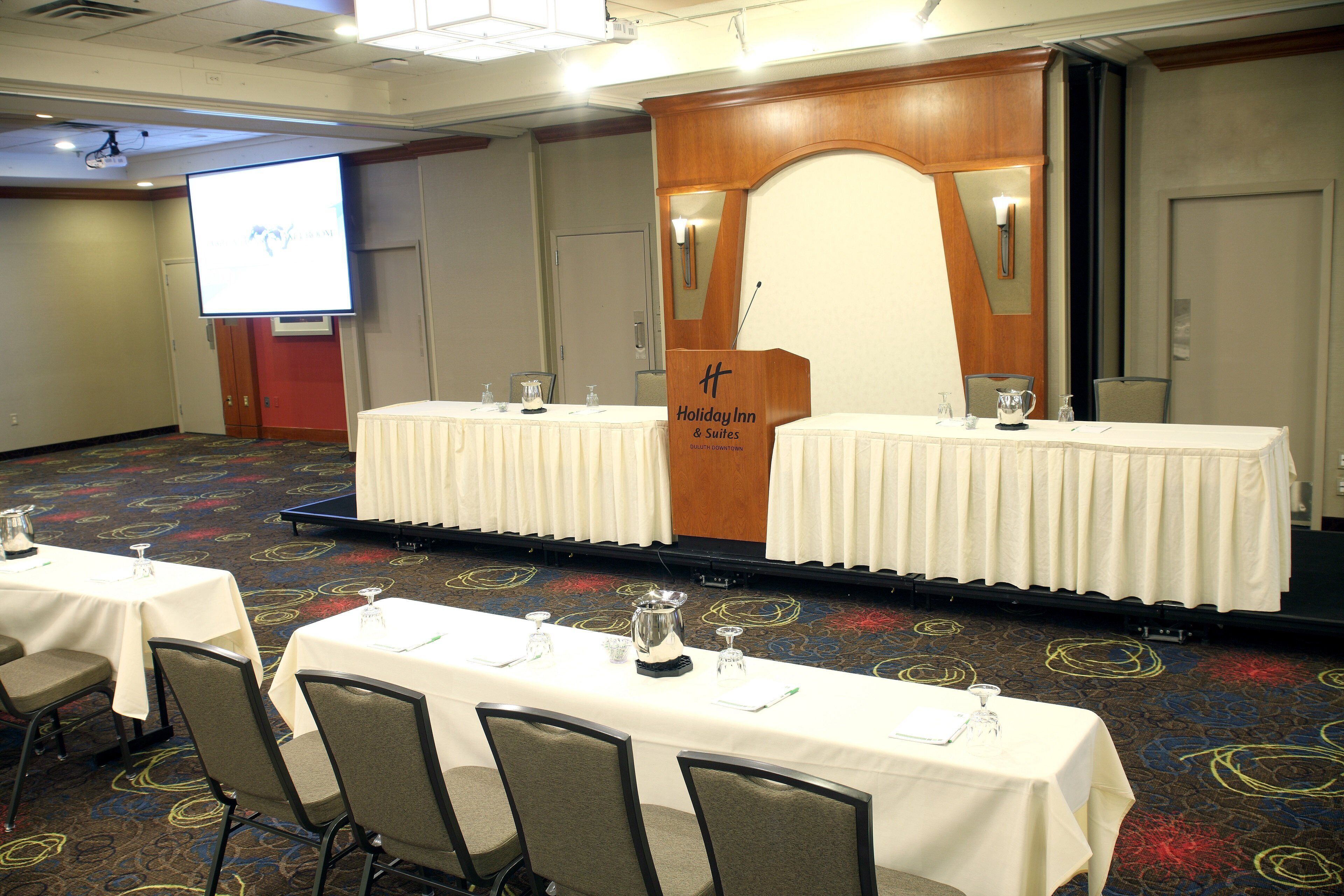 Un salón de conferencias con dos mesas, sillas, un atril y una pantalla de proyección.