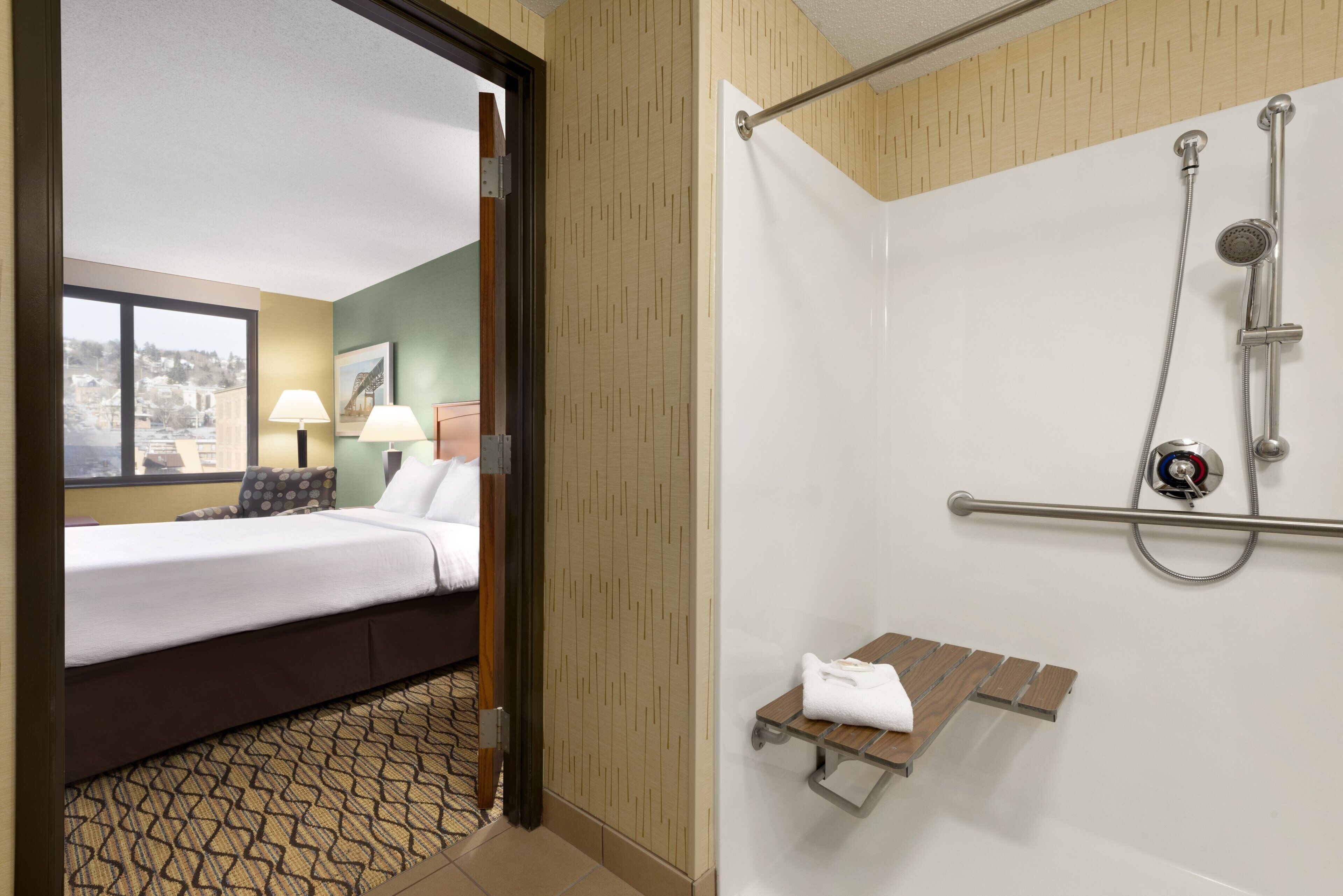 A hotel room with a bed, a window with a view, a shower with a handrail, and a towel rack.