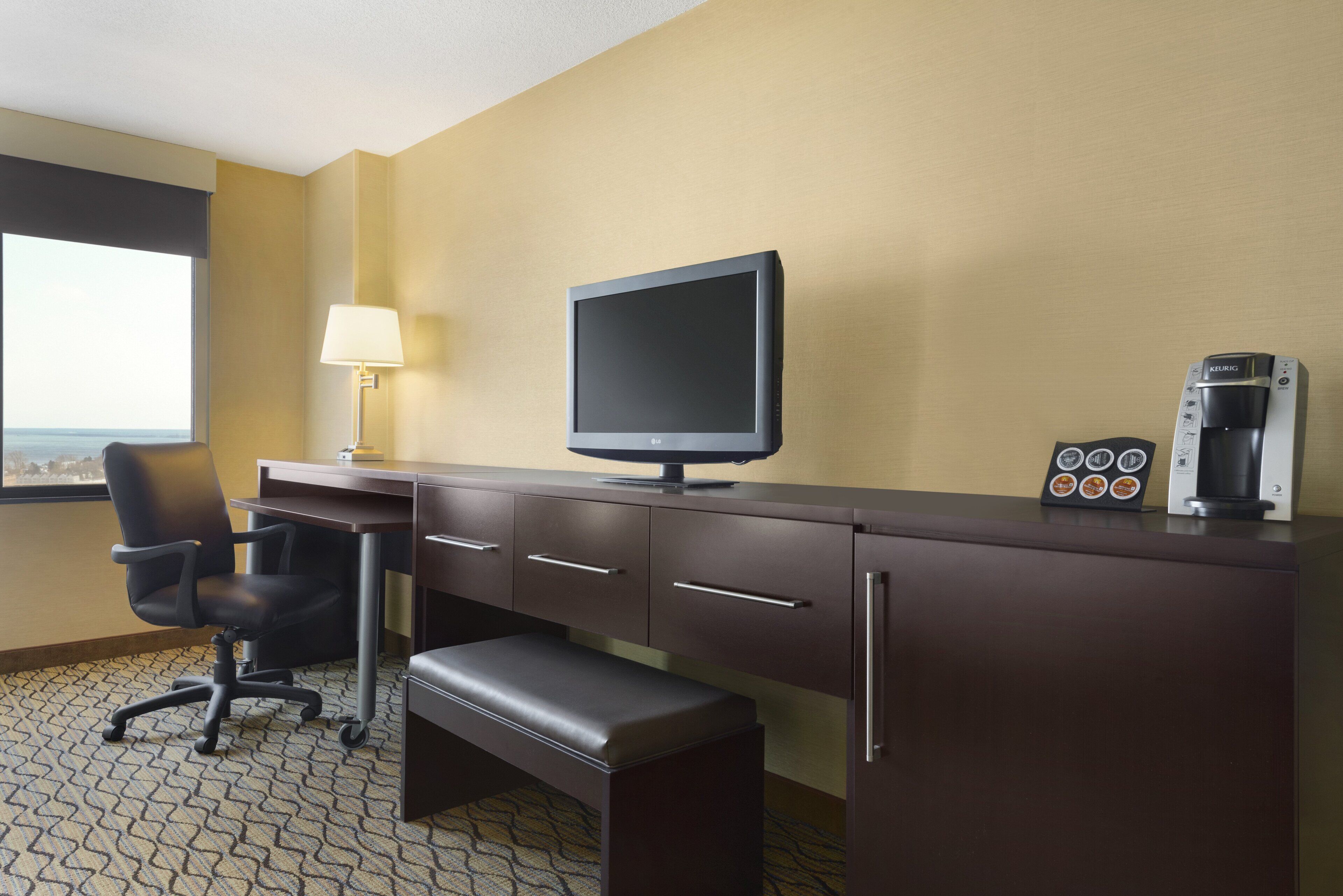 A hotel room with a desk, a chair, a flat-screen TV, a coffee maker, and a window with a view.
