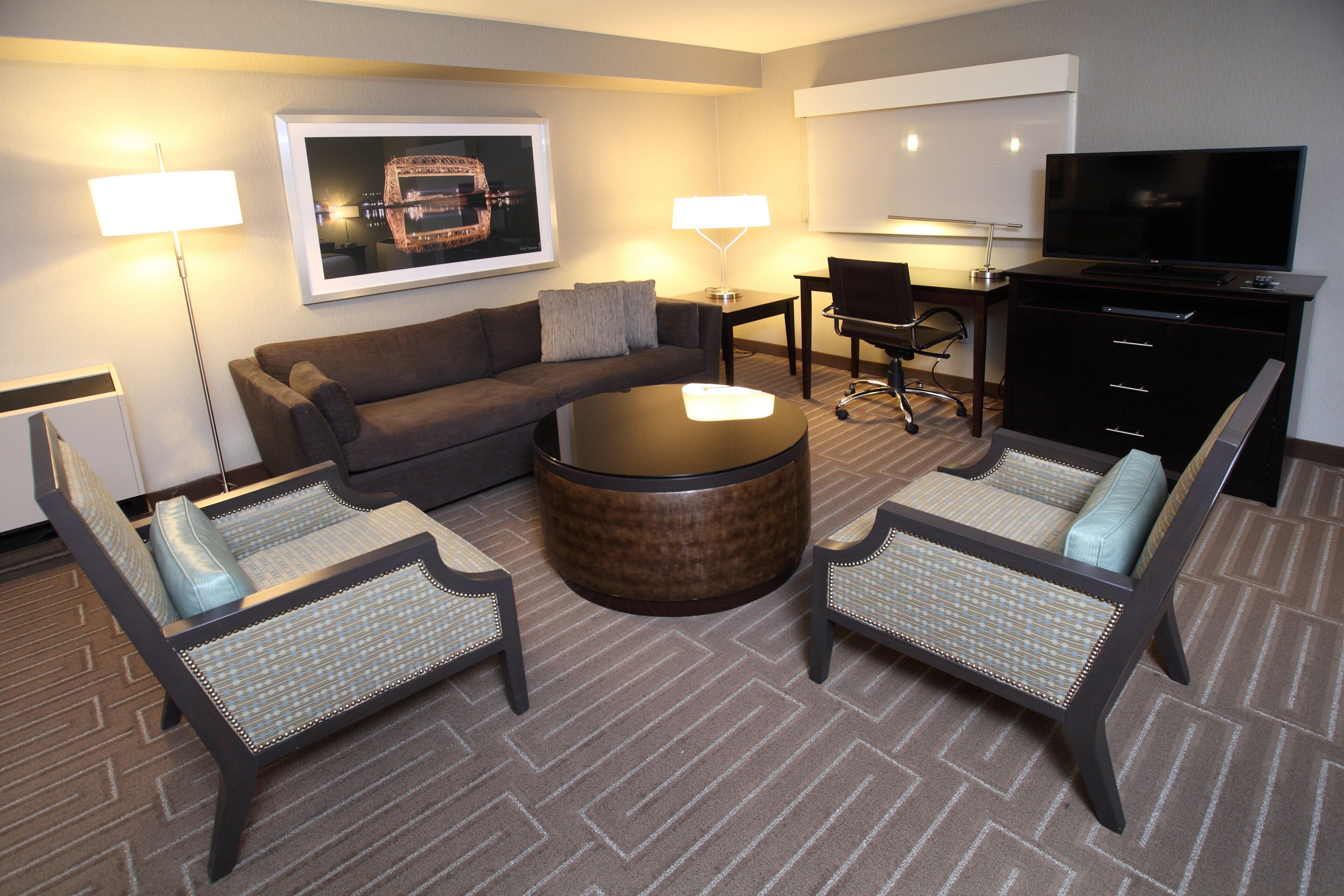 A hotel room with a sofa, two armchairs, a coffee table, a desk with a chair, a TV, and a framed picture on the wall.