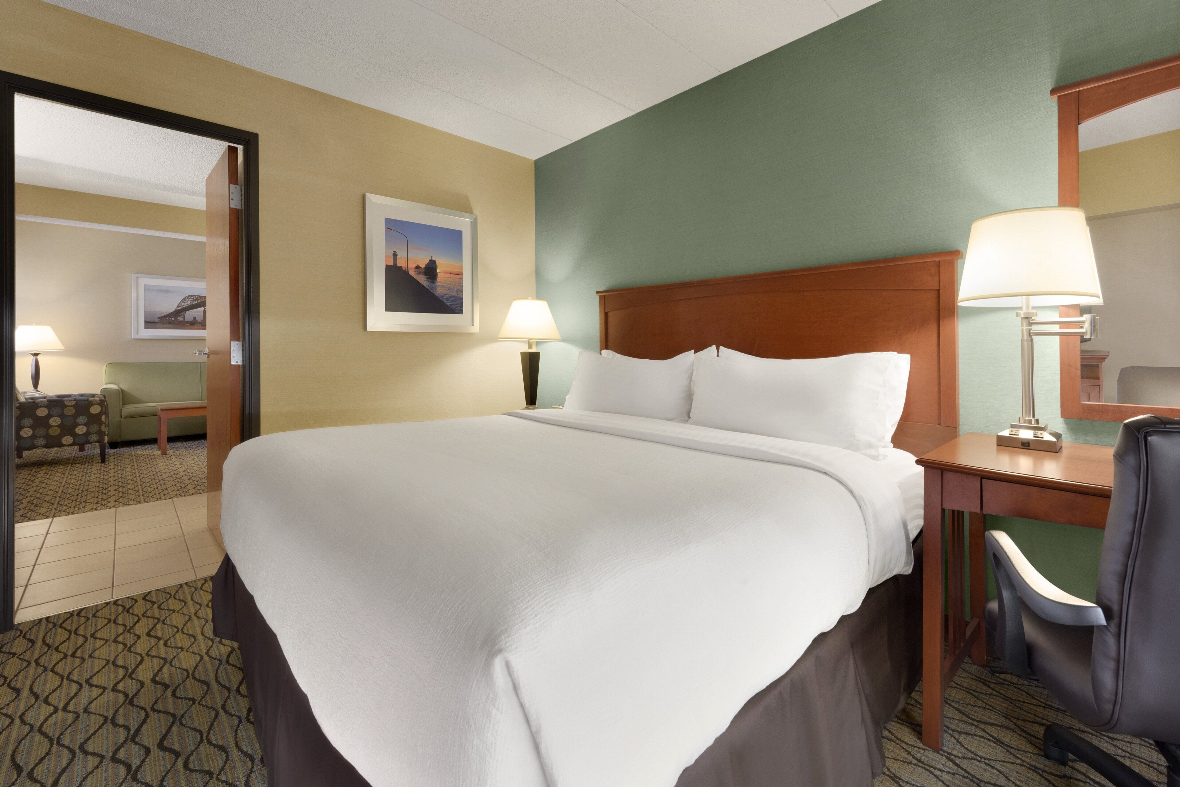 Una camera d'albergo con un letto grande, due comodini con lampade, una sedia, una scrivania e un quadro appeso al muro.