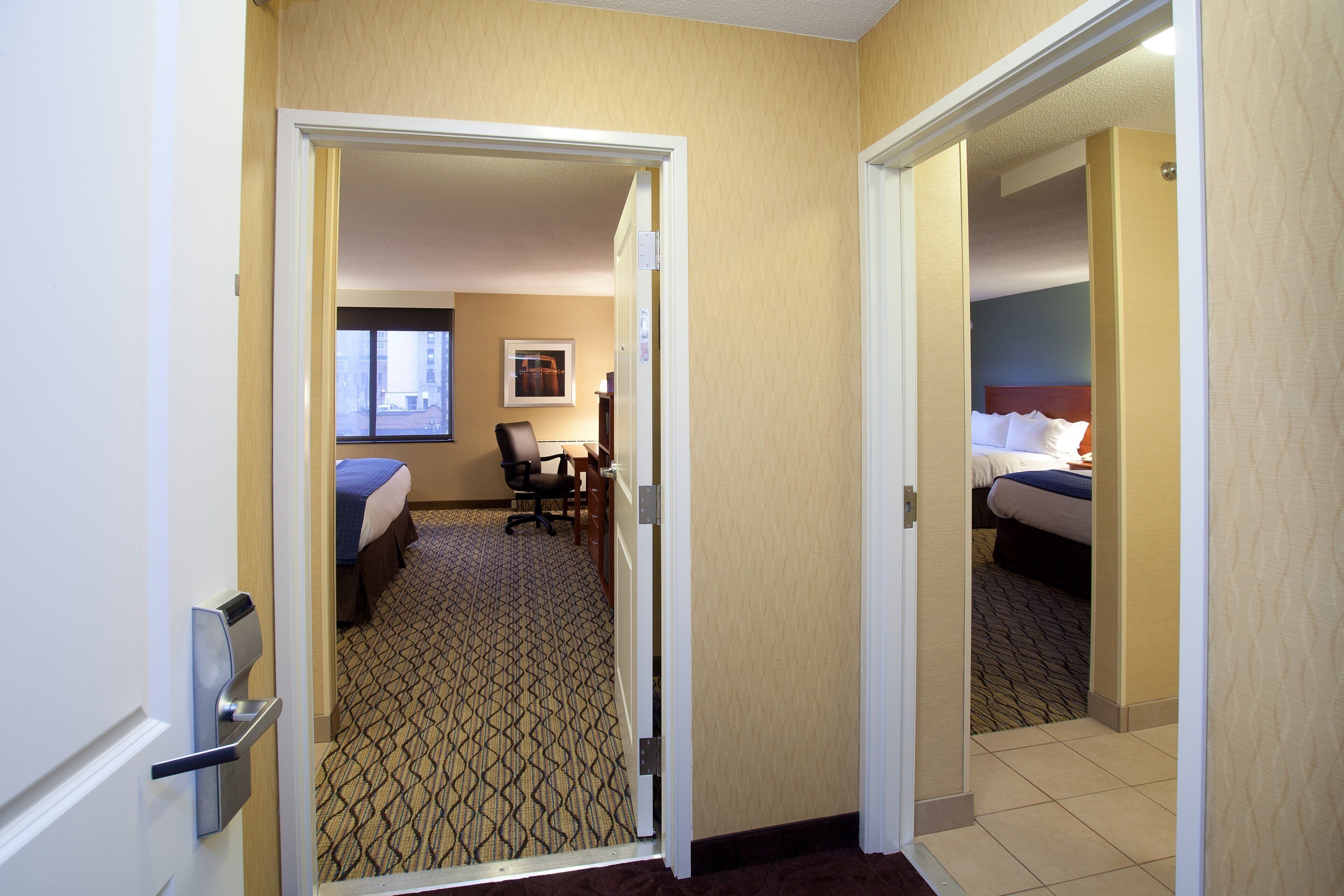 Ein Hotelzimmer mit einem Bett, einem Schreibtisch und einem Sessel. Ein Fenster bietet Blick auf die Gebäude außerhalb.