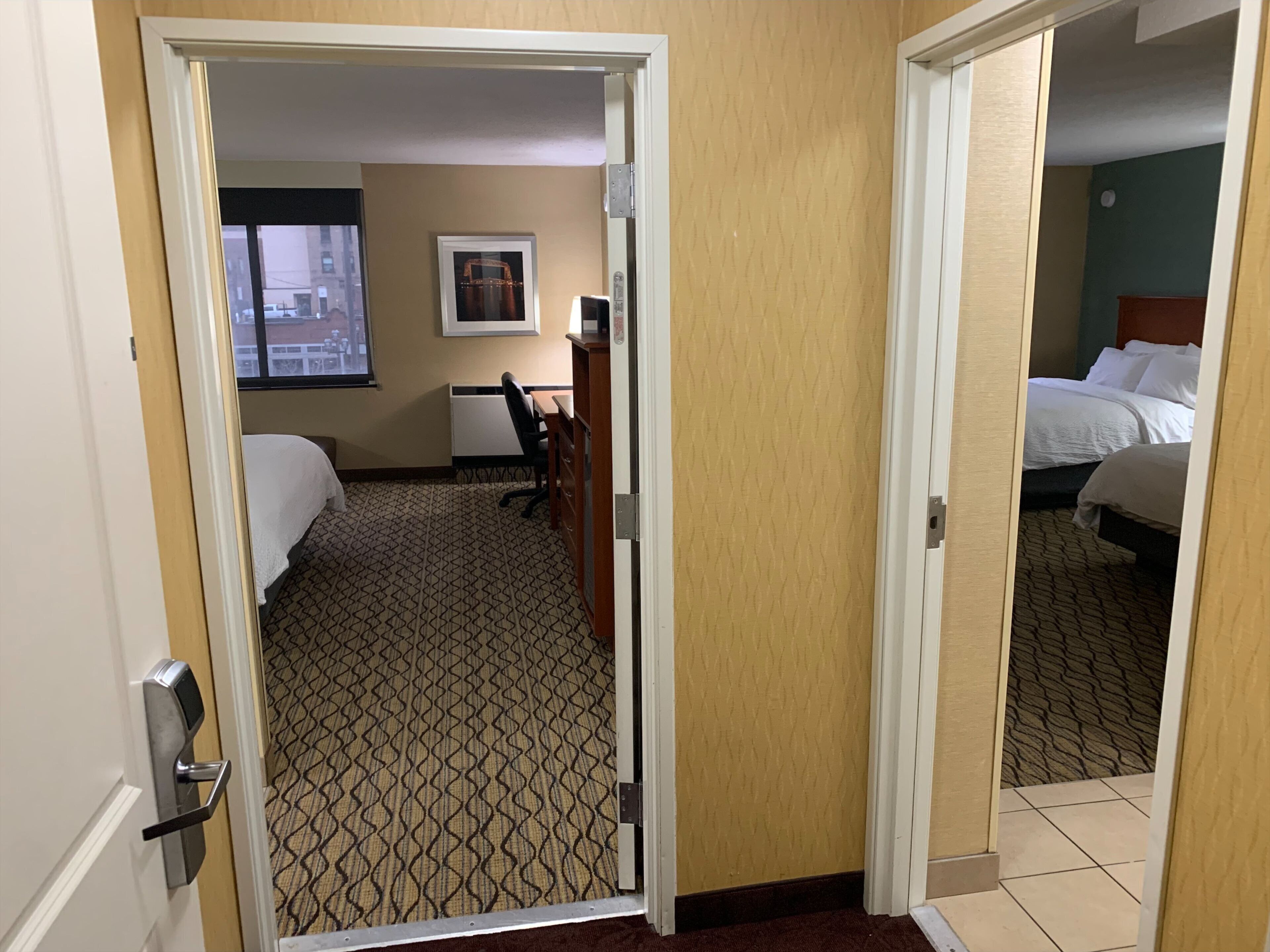A hotel room with a patterned carpet, a desk with a chair, a bed, and a window with a view of buildings.