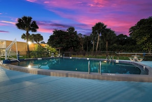 A swimming pool with a surrounding fence, palm trees, and a sunset sky. - HomeTowne Studios By Red Roof Vero Beach - I-95 (Vero Beach)