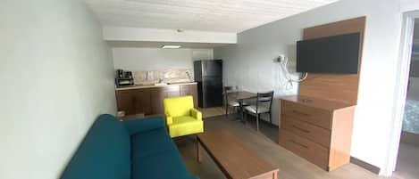 A compact living space with a sofa, a coffee table, a dining table with chairs, a kitchen area, and a flat-screen TV mounted on the wall.