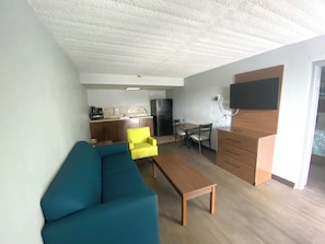 A compact living space with a sofa, a coffee table, a dining table with chairs, a kitchen area, and a flat-screen TV mounted on the wall. - HomeTowne Studios By Red Roof Vero Beach - I-95 (Vero Beach)