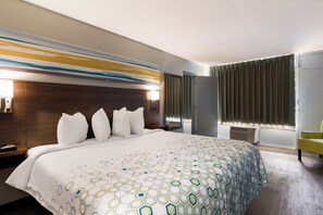 A hotel room with a large bed, a wooden headboard, a wall-mounted lamp, a window with curtains, and a bedside table with a lamp. - HomeTowne Studios By Red Roof Vero Beach - I-95 (Vero Beach)
