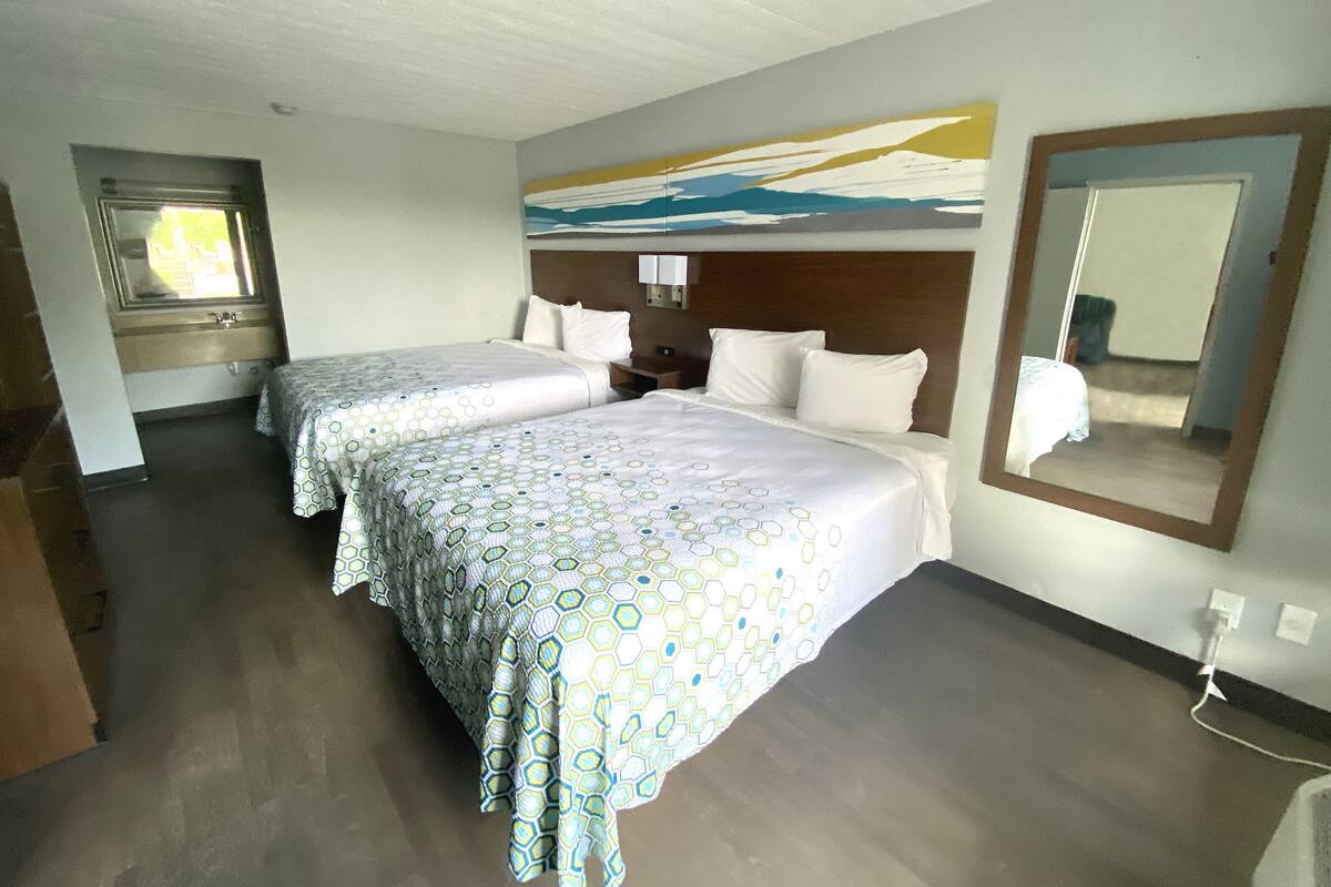 A hotel room with two beds, a large window, a painting on the wall, and a mirror.