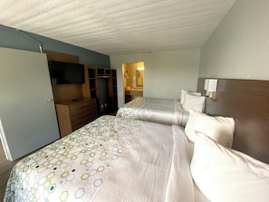 A hotel room with two beds, a TV, a wardrobe, and a bathroom visible in the background.