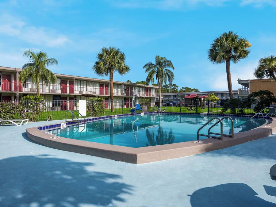 A swimming pool with a blue tiled edge, surrounded by lounge chairs, palm trees, and a hotel building with red doors and balconies.