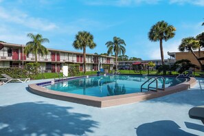 A swimming pool with a blue border, surrounded by lounge chairs and palm trees. - HomeTowne Studios By Red Roof Vero Beach - I-95 (Vero Beach)
