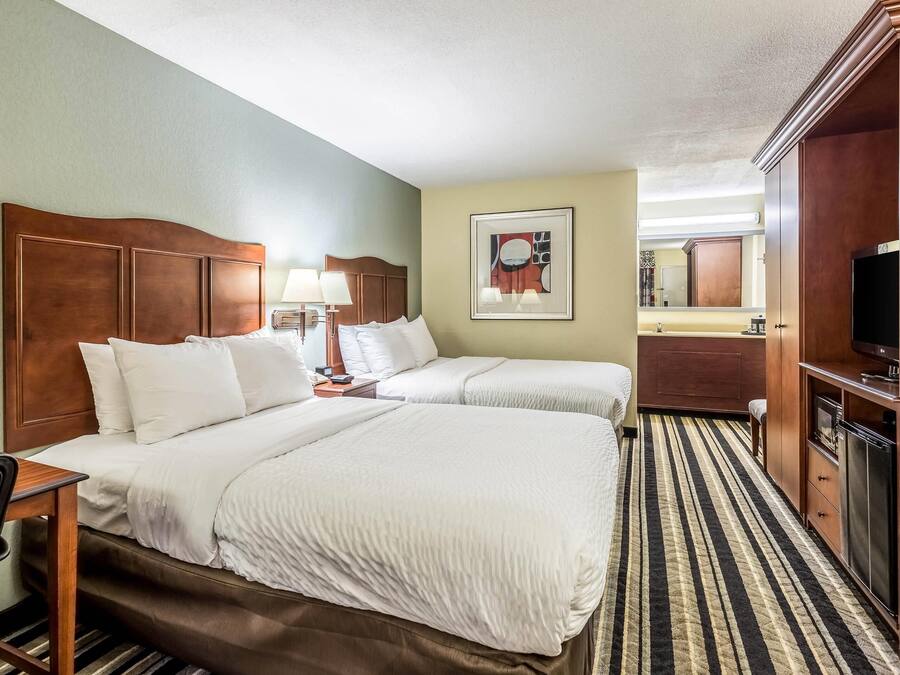 A hotel room with two beds, a desk, a chair, a television, and a bathroom with a mirror.