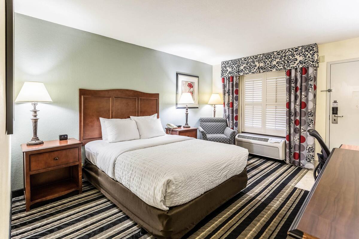A hotel room with a large bed, two bedside tables with lamps, a chair, a window with curtains, and a radiator.