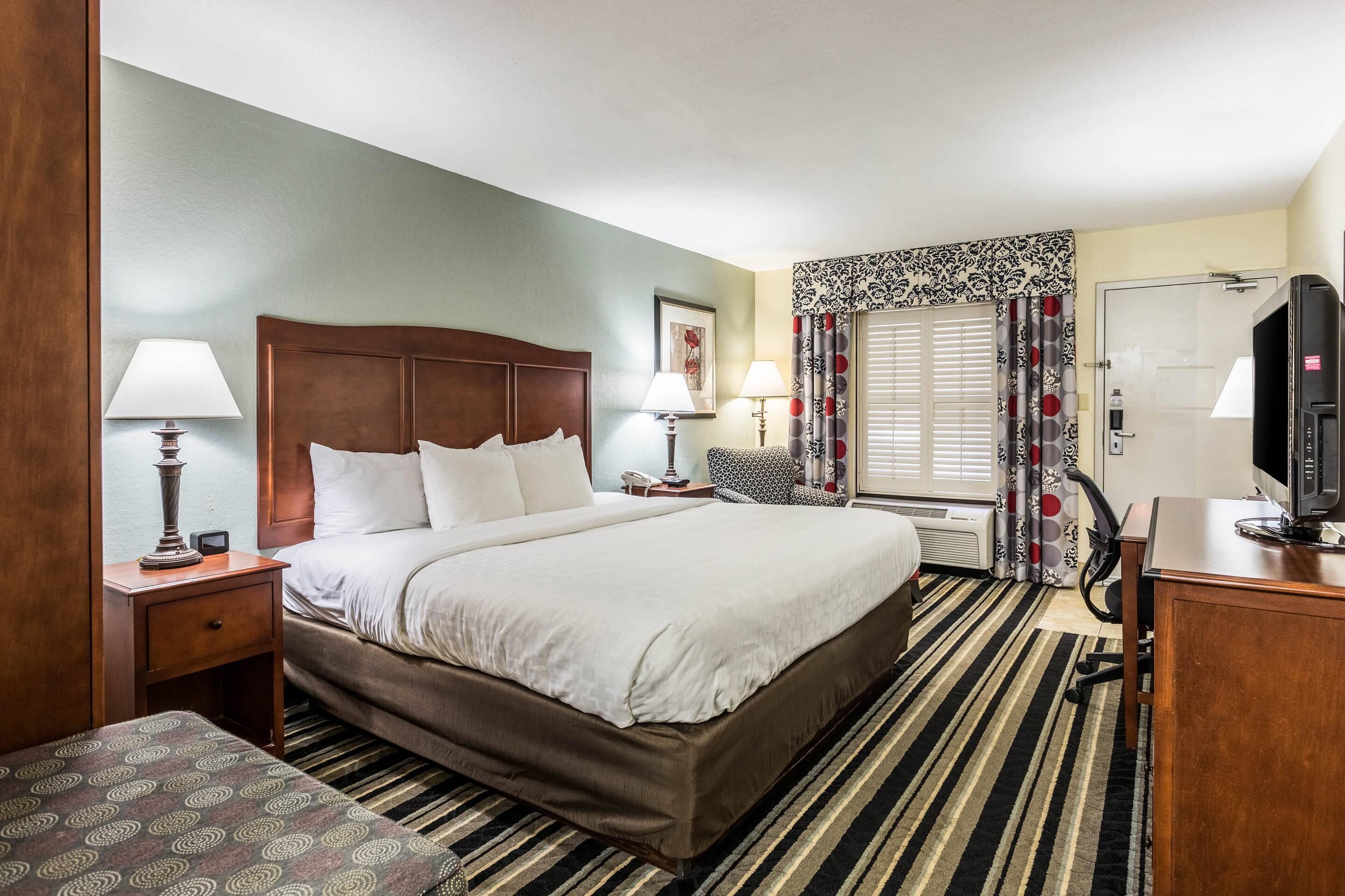 A hotel room with a large bed, two bedside tables with lamps, a desk with a computer, a window with patterned curtains, and a door with a glass panel.