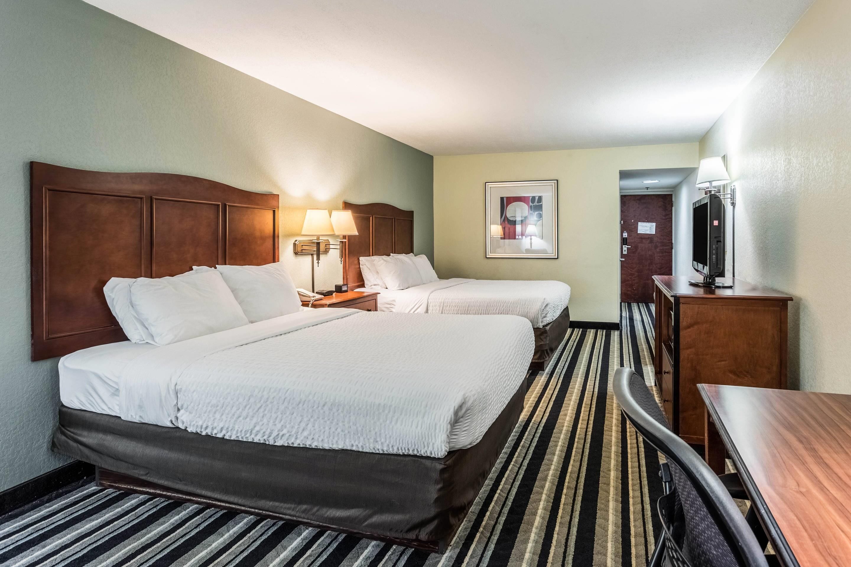 A hotel room with two beds, a desk, a chair, a TV, and a lamp.